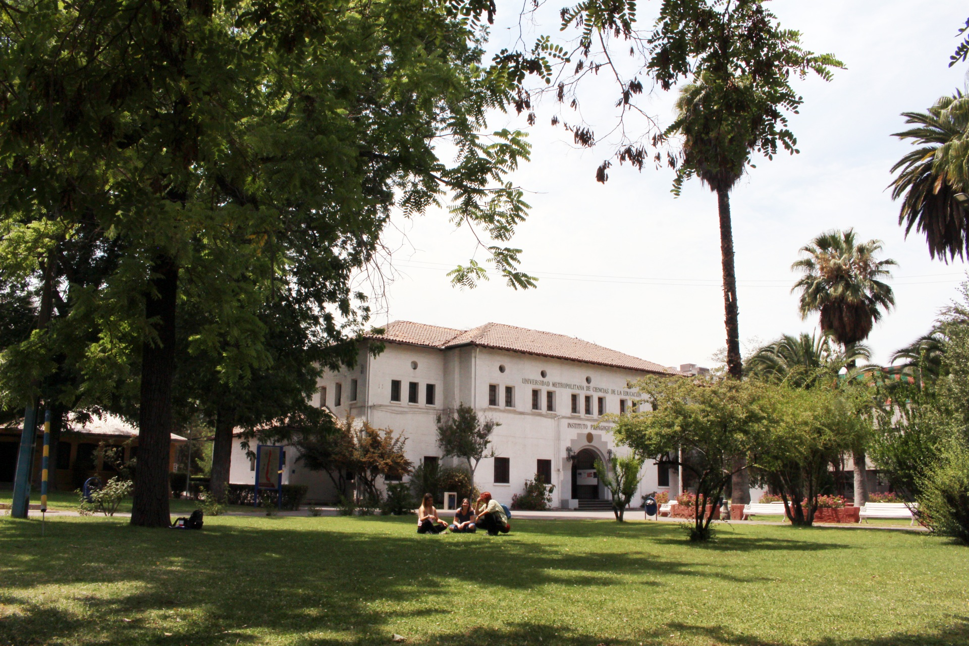 UMCE - Una edificio de estilo colonial con un jardín amplio, donde varias personas están sentadas en el césped disfrutando del sol. Árboles y palmeras rodean el área, creando un ambiente natural.