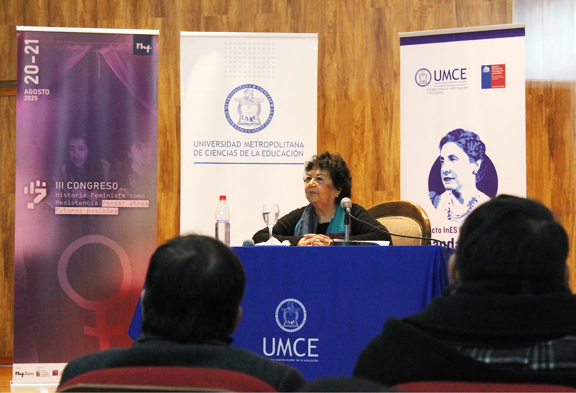 Académica exponiendo en el III Congreso “Historia Feminista como Resistencia: Pensar otros futuros posibles”, realizado en la Universidad Metropolitana de Ciencias de la Educación (UMCE). En la mesa se observa un micrófono, agua y el logo institucional, con público asistente en primer plano.