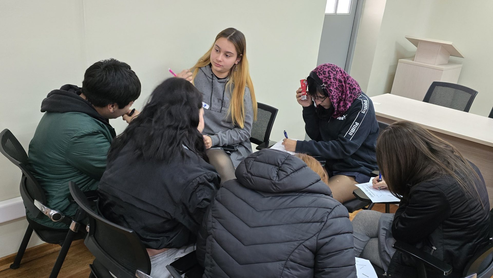 Un pequeño grupo de personas sentadas en sillas, conversando sentados con cuadernos y lápices, rodeados de una habitación de color claro.