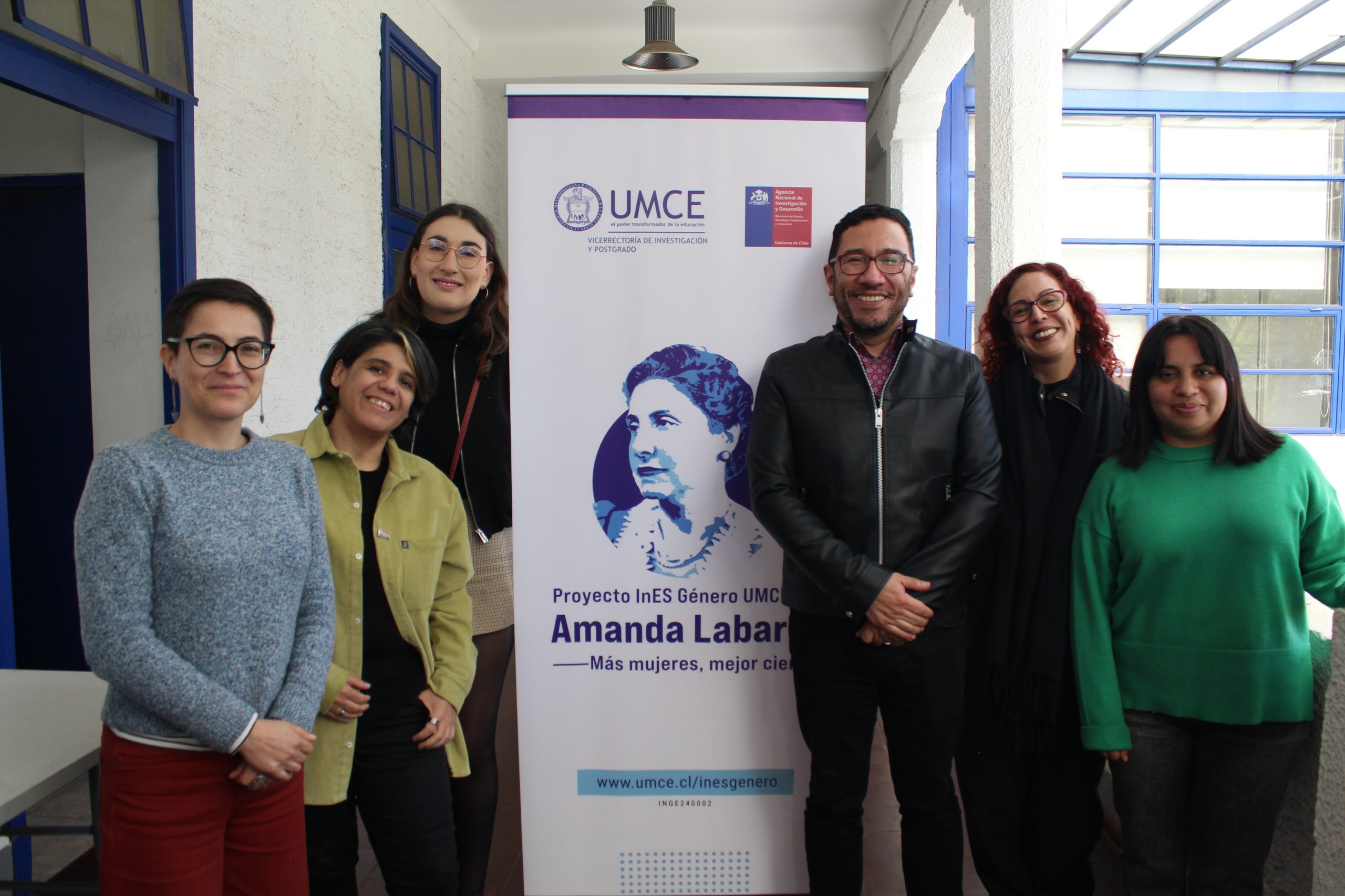 Fotografía del equipo InES Género junto a personas invitadas al conversatorio paradas sonriendo a cámara junto a pendón que dice Proyecto InES Género UMCE Amanda Labarca. Más mujeres, mejor ciencia
